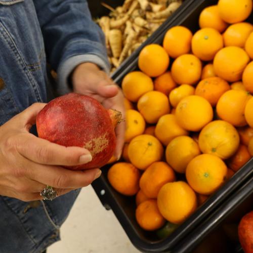 Rhory holding produce at ACBC Food Shelf A woman's hands holding a pomegranate in front of a rack of oranges at ACBC Food Shelf