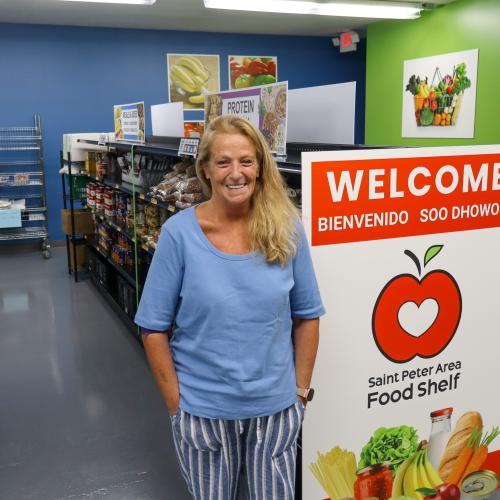 Jodi in St. Peter Area Food Shelf woman in blue shirt standing beside a welcome sign next shelves of food in food shelf