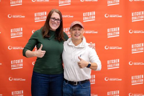 A woman in green and a man in a red hat pose in front of a red KitchCo mural